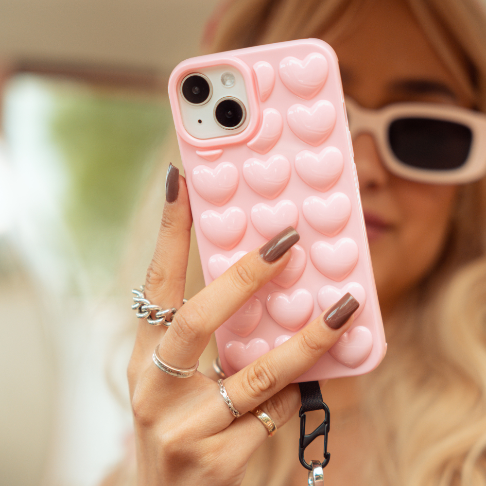 A person with wavy blonde hair and white sunglasses holds a Coconut Lane 3D Hearts Phone Case in Candyfloss Pink. The individual has manicured nails adorned with multiple rings, set against a blurred indoor background.
