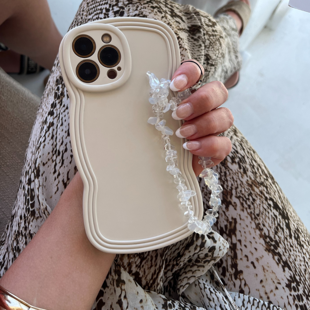 A person holds a beige smartphone with the NAKD Curvy Phone Case - Vanilla by Coconut Lane, featuring a unique wavy-edged design and paired with a clear beaded strap. The person's nails are manicured in light pink, and they are wearing a snake-print patterned outfit. A gold bracelet adorns their wrist, perfectly complementing the highly protective yet lightweight phone case from Coconut Lane.
