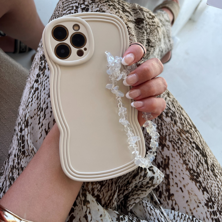 A person holds a beige smartphone with the NAKD Curvy Phone Case - Vanilla by Coconut Lane, featuring a unique wavy-edged design and paired with a clear beaded strap. The person's nails are manicured in light pink, and they are wearing a snake-print patterned outfit. A gold bracelet adorns their wrist, perfectly complementing the highly protective yet lightweight phone case from Coconut Lane.