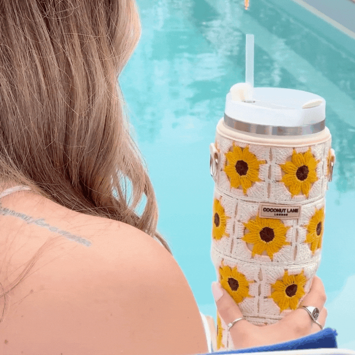 A person with long, wavy hair, seen from behind, holds a Coconut Lane Tumbler Carry Case - Sunflower Crochet. The person is by a pool, with one shoulder visible, wearing a sleeveless top. The water is bright blue and the overall scene has a relaxed, summery vibe.