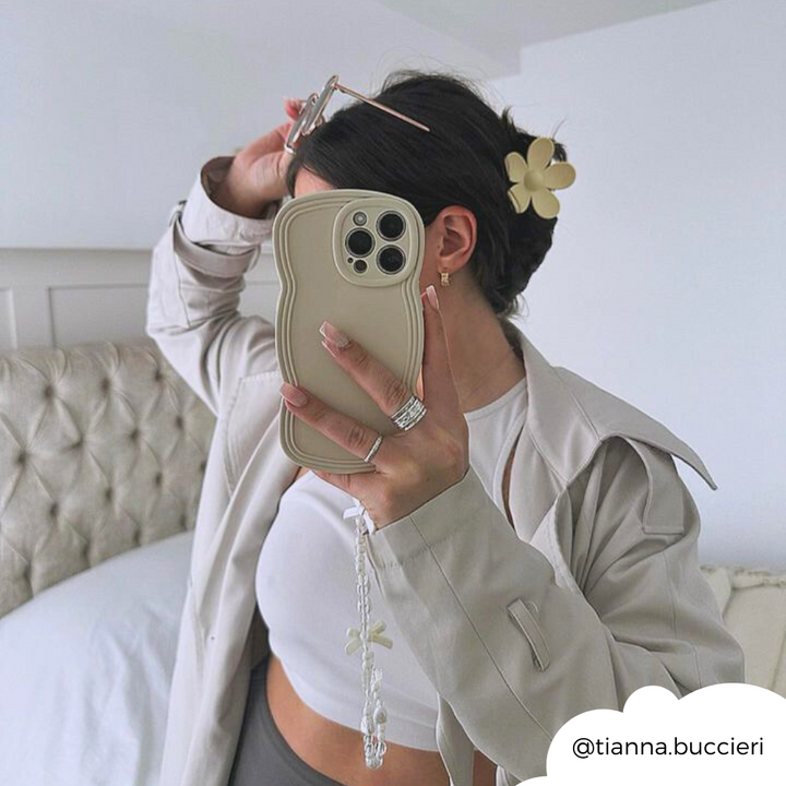 Person takes a mirror selfie with an iPhone in a highly protective, beige wavy case by Coconut Lane's NAKD Curvy Phone Case in Vanilla. They're wearing a cream jacket and white crop top, their hair is up with a flower clip. A white bed and minimal decor are in the background. Instagram handle "@tianna.buccieri" is displayed.