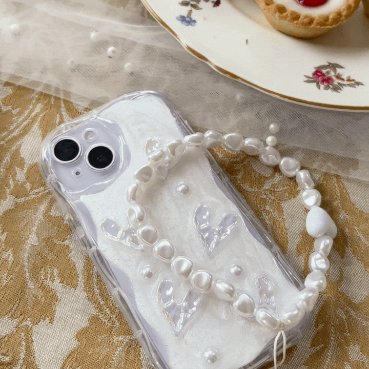 A white iPhone with a clear case featuring white heart designs and pearl accents lies on a beige, lace-like fabric. Draped over the phone is a delicate pearl bracelet. Attached is the Coconut Lane Beaded Phone Strap - Chunky Pearls, perfectly complementing the design. Nearby, a white floral plate holds a delicious pastry.
