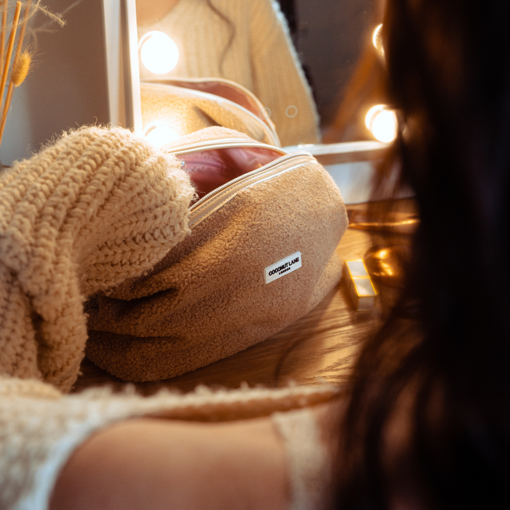 Person in a cozy sweater at a vanity with a beige, teddy-textured Coconut Lane expandable makeup bag and a lit mirror.
