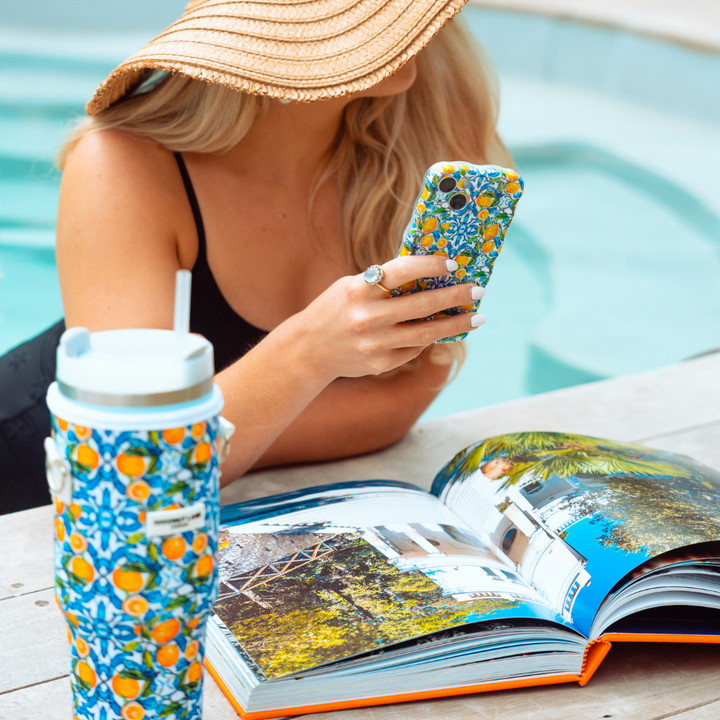 A person with long blonde hair and a sunhat, wearing a black swimsuit, lounges by a pool. They are looking at a smartphone encased in the extra protective Wavy Phone Case - Amalfi Lemon from Coconut Lane. In front of them is an open book and a large, patterned drink tumbler.