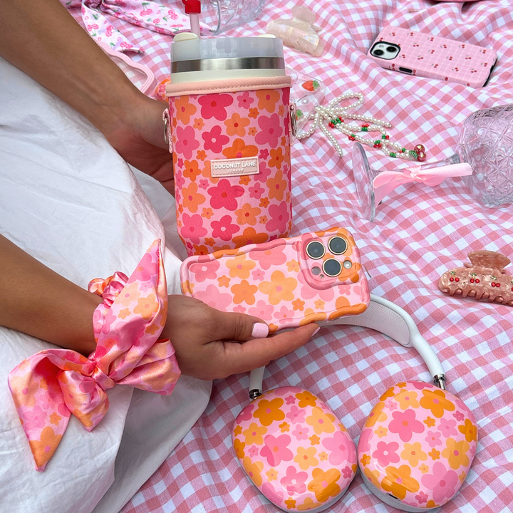 A person is holding an iPhone with a pink and orange floral case, perfectly matching their Coconut Lane Tumbler Carry Case - Pink & Orange Bloom. Also seen is a hair scrunchie, headphones, and various accessories laid out on a pink and white checkered picnic blanket.