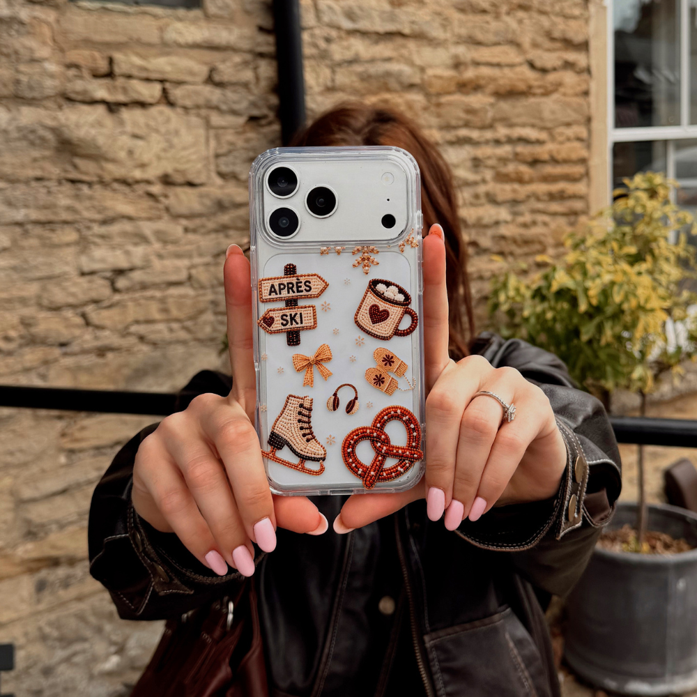 A person holds up a clear phone case with winter and coffee-themed decorations , including a heart coffee mug, pretzel, ice skate, and sign that says 'APRÈS / SKI'. The background shows a stone wall and outdoor plants.