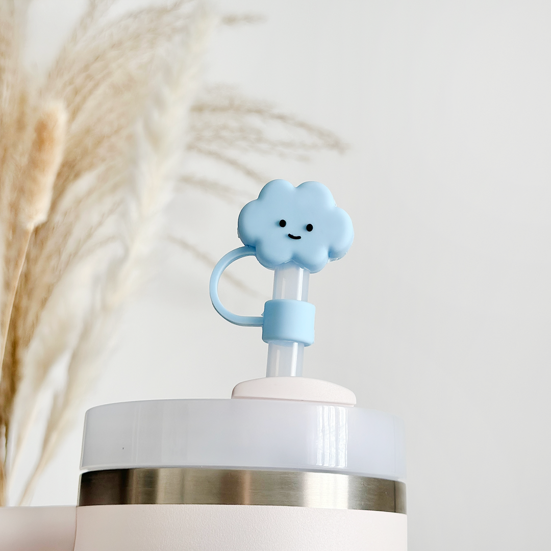 The close-up image showcases the Blue Cloud Straw Cover by Coconut Lane, a light blue, cloud-shaped straw topper made of high-quality silicone with a cheerful smiley face. It sits atop a cup with a transparent lid accented by a metal band. The background features softly-focused, wheat-like plants set against a neutral pastel backdrop.
