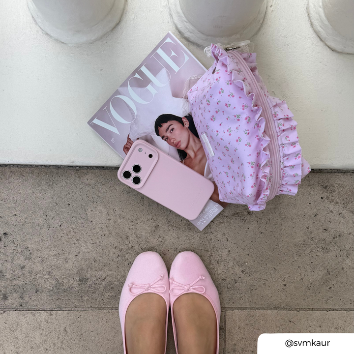 A styled flat-lay featuring a light pink floral ruffled zippered makeup bag, a baby pink phone case, and a Vogue magazine on a concrete surface, with a person's feet in pink ballet flats visible at the bottom.