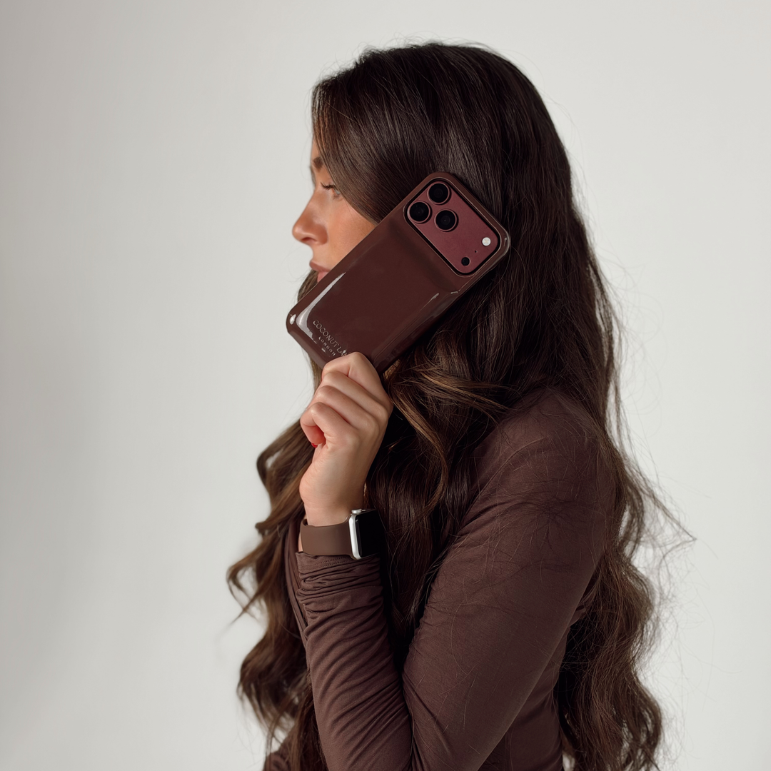 Woman wearing a brown pilates outfit, holding an iphone up to her ear. The phone is in a glossy chocolate brown Coconut Lane puffer phone case with embossed logo. On her wrist is a Coconut Lane NAKD Chocolate brown apple watch strap.
