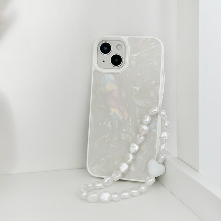 A white smartphone with a shimmering, iridescent case is propped upright against a white corner. The phone case boasts Coconut Lane's stylish Beaded Phone Strap - Chunky Pearls, adorned with irregularly shaped white pearls and ending in a small white heart. The background and surface are also white, creating a minimalist aesthetic.