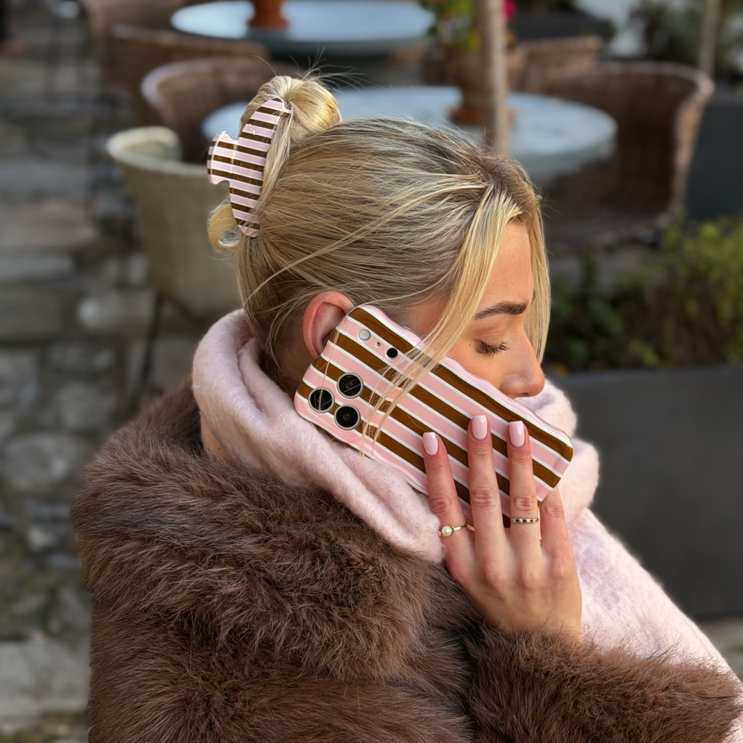 Woman with blonde hair. In her hair is a cocoa and pink stripe Claw Clip featuring glossy chocolate brown, blush pink and cream vertical stripes. In her hand is the cocoa and pink stripe wavy phone case. 