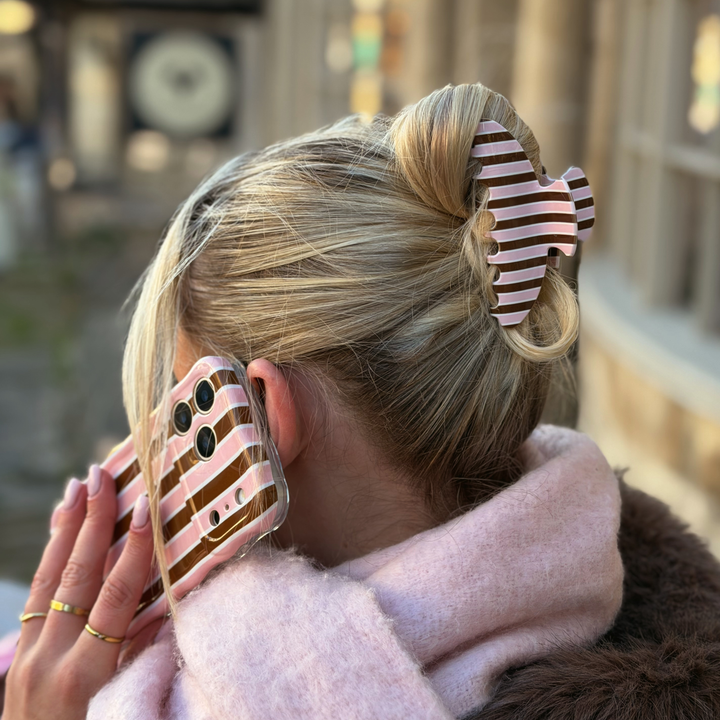 Woman with blonde hair. In her hair is a cocoa and pink stripe Claw Clip featuring glossy chocolate brown, blush pink and cream vertical stripes. In her hand is the cocoa and pink stripe wavy phone case. 