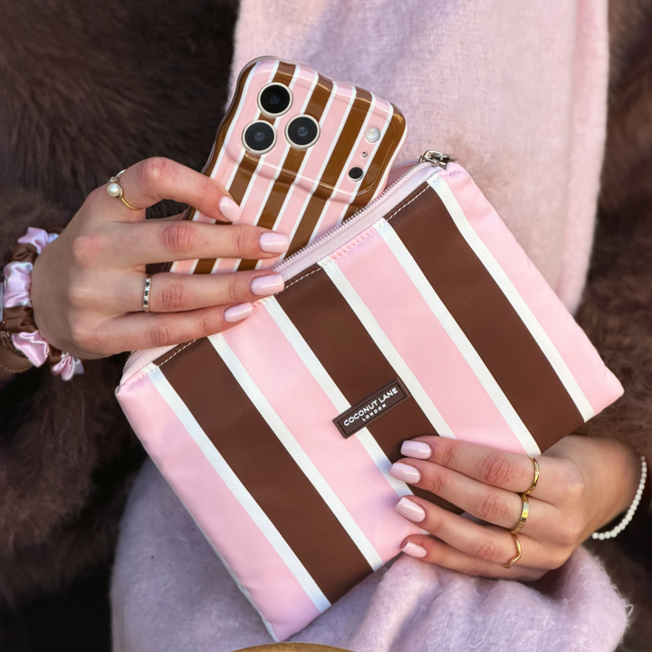 Woman holding the Coconut Lane Cocoa & Pink Stripe Everything Pouch featuring chocolate brown, pastel pink and white vertical stripes. She is pulling out the matching wavy phone case. On her wrist is the matching satin scrunchie. She is wearing a baby pink scarf and a faux fur brown jacket.
