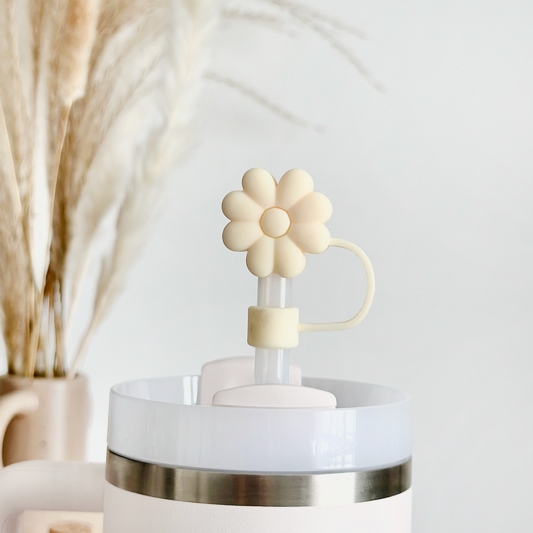 A close-up of a white insulated tumbler with a large plastic straw, adorned with Coconut Lane's Cream Flower Straw Cover - a silicone daisy-shaped topper. In the background, dried pampas grass arranged in a beige vase enhances the natural aesthetic of the scene.