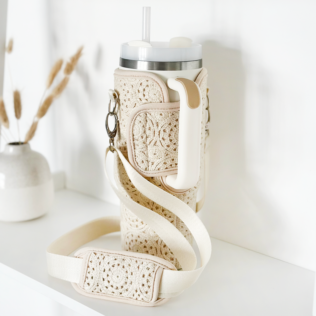 A tall, beige tumbler covered in Coconut Lane's Festival Crochet Tumbler Carry Case with a handle sits on a white surface. It features a white strap for easy carrying. In the background, a minimalist white vase with dried grass stems adds to the clean, simple aesthetic.