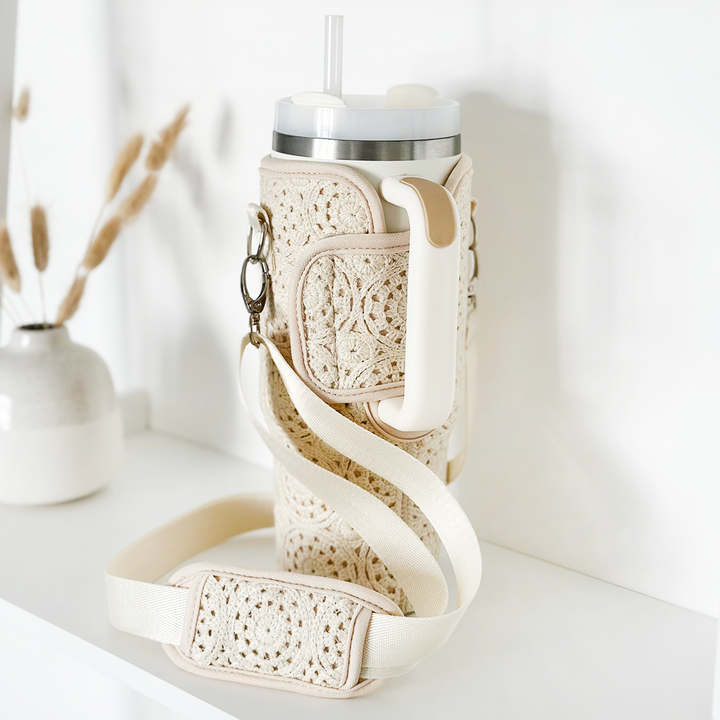 A tall, beige tumbler covered in Coconut Lane's Festival Crochet Tumbler Carry Case with a handle sits on a white surface. It features a white strap for easy carrying. In the background, a minimalist white vase with dried grass stems adds to the clean, simple aesthetic.