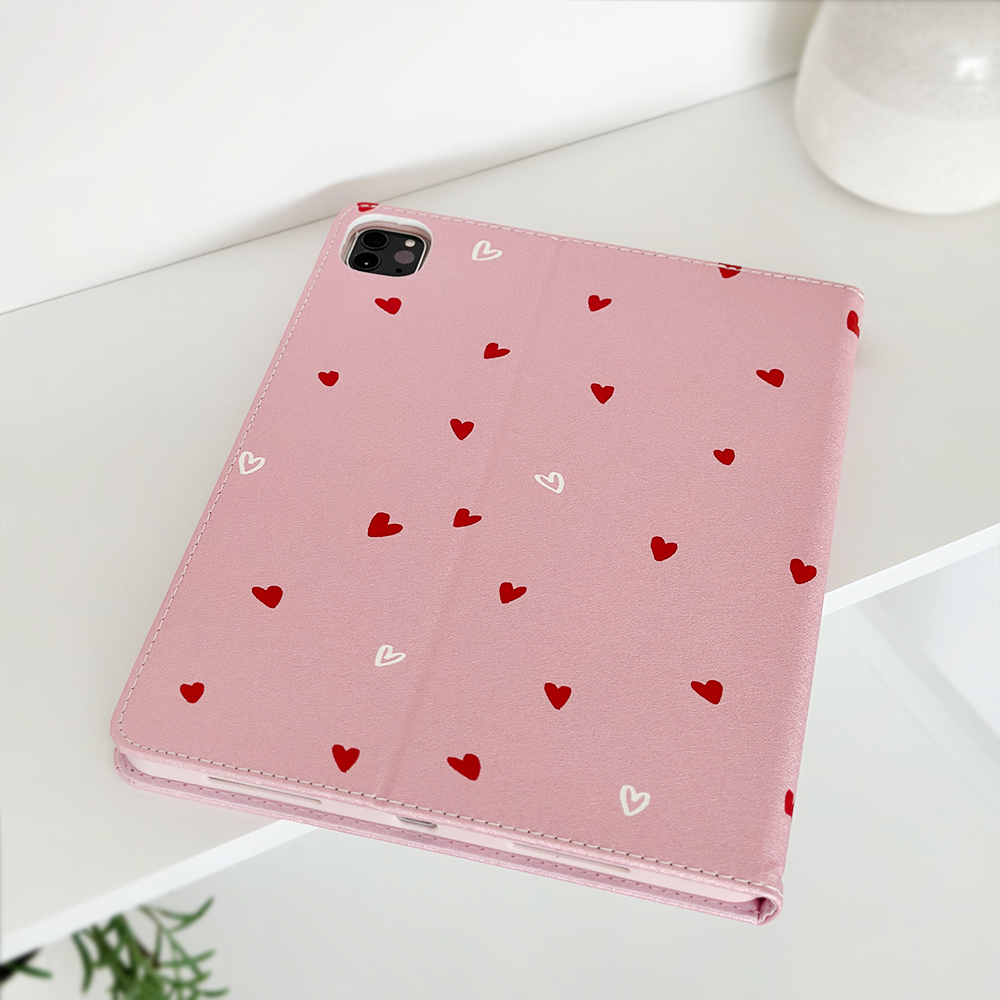 A pink Coconut Lane Cute Hearts iPad Case, adorned with a charming pattern of small red and white hearts, is displayed on a white surface. The open camera slot clearly suggests compatibility with a device that has a rear camera. A hint of a plant can be seen in the lower left corner, accompanied by a white ceramic vase in the upper right corner.