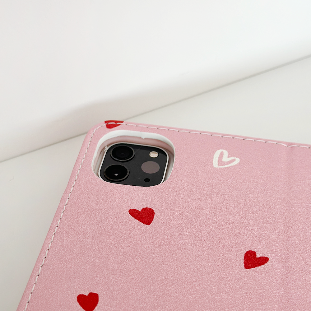 A close-up image of a Cute Hearts iPad Case by Coconut Lane, featuring a pink PU Vegan Leather design adorned with an adorable heart pattern near the camera area of an iPad. The case is laid flat on a white surface.