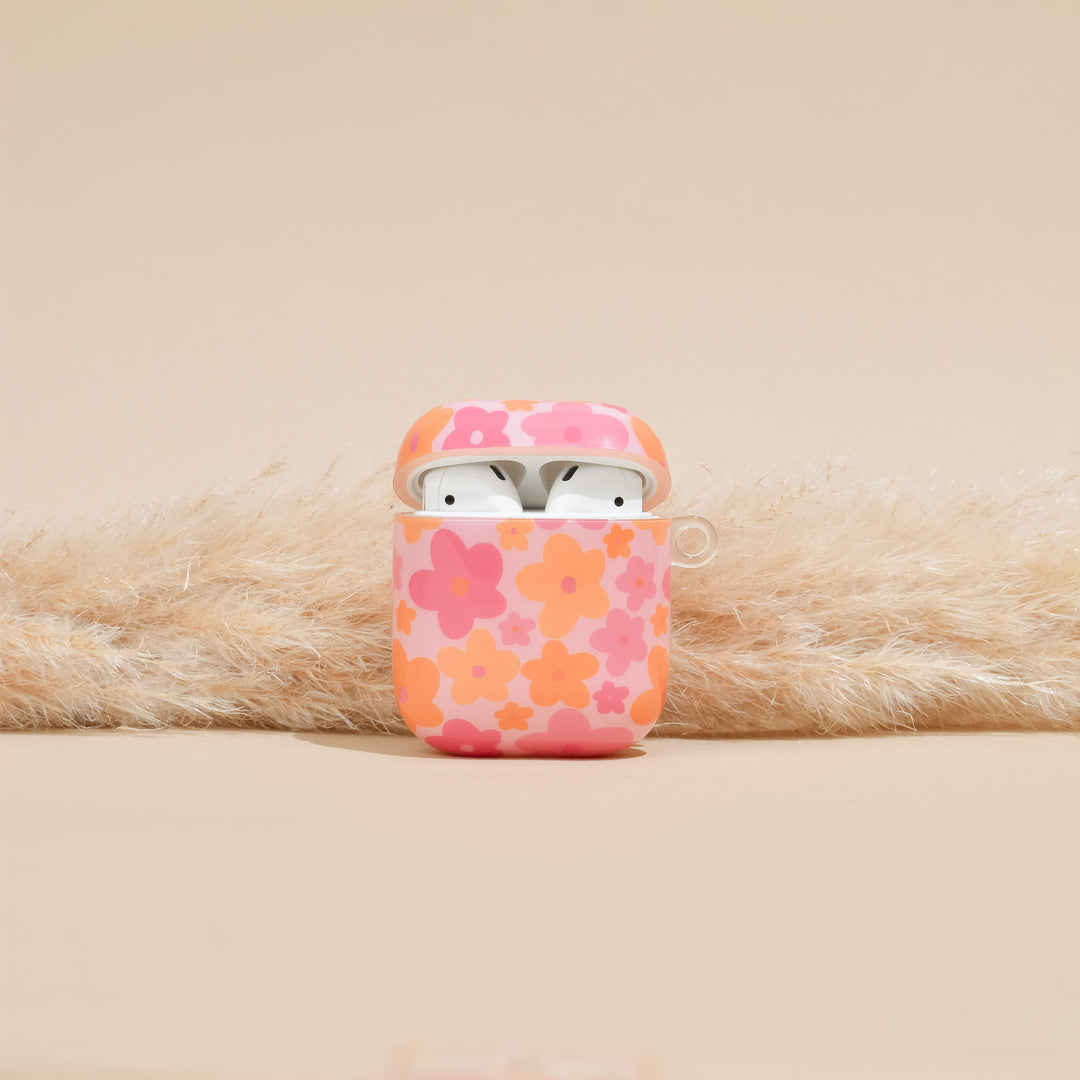 The Coconut Lane AirPods Case - Pink & Orange Bloom is displayed against a beige background, housing a pair of white wireless earbuds. Utilizing IMD Print Technology, the vibrant and durable floral design enhances the case's appeal. The lid is open, revealing the earbuds inside, while pampas grass arranged behind adds a decorative touch.
