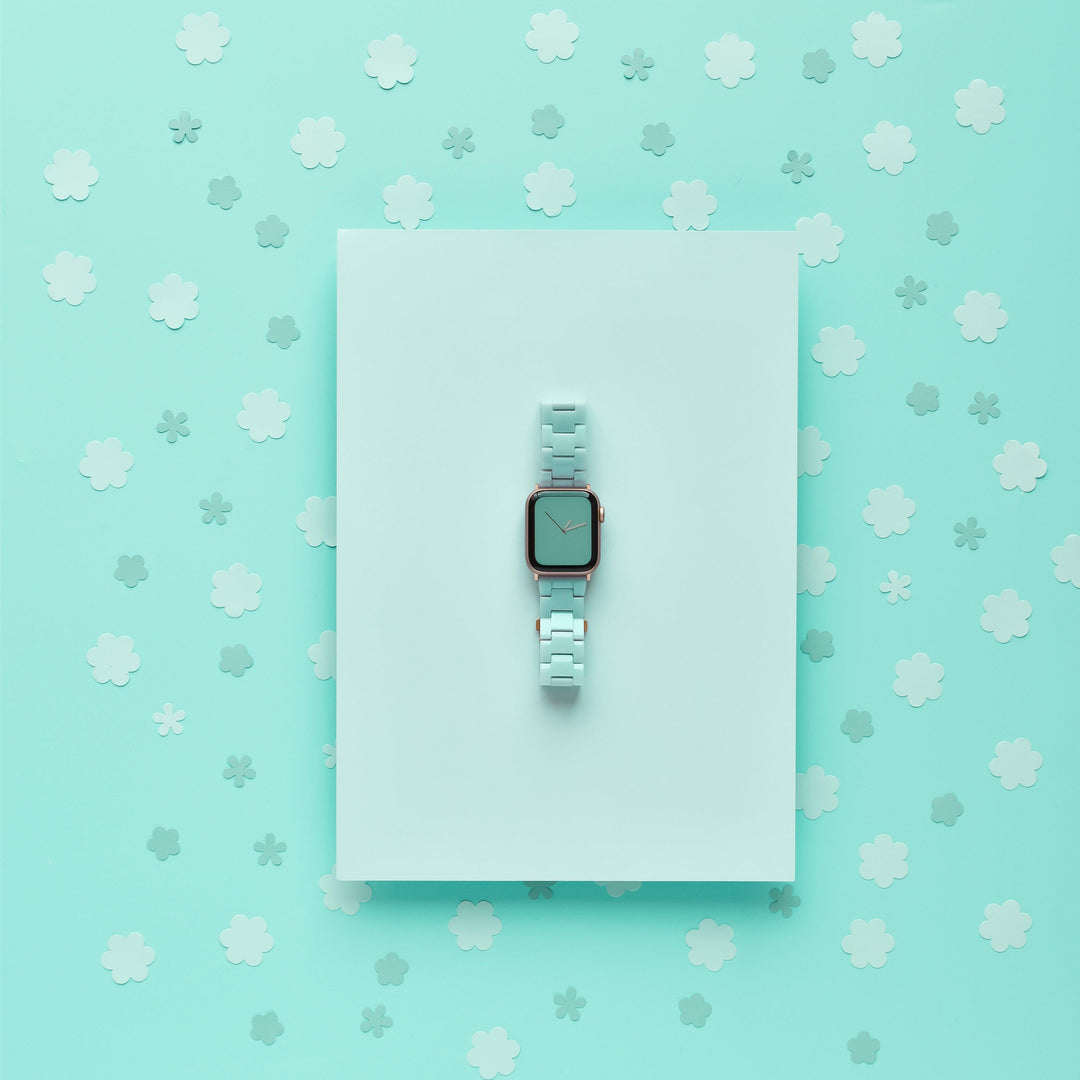 A minimalist photo of a light blue smartwatch featuring the Luxe Pastel Mint Apple Watch Strap by Coconut Lane, centered on a light blue rectangular surface. The background is a similar shade of blue, adorned with numerous small, white, cloud-like shapes scattered around.