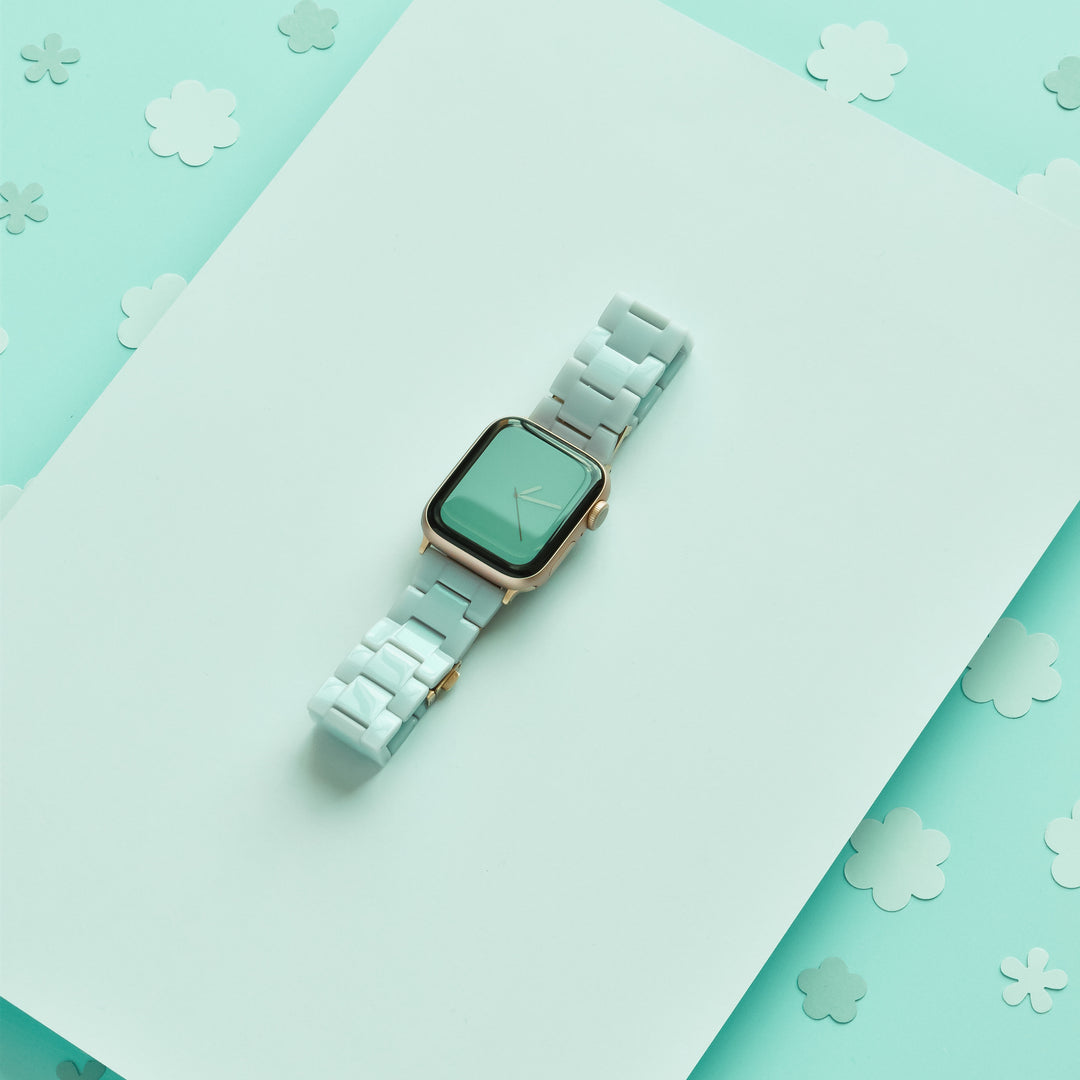 The Luxe Pastel Mint Apple Watch Strap by Coconut Lane sits elegantly on a light teal backdrop adorned with scattered flowers in various shades of teal and white.