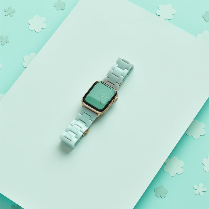 The Luxe Pastel Mint Apple Watch Strap by Coconut Lane sits elegantly on a light teal backdrop adorned with scattered flowers in various shades of teal and white.