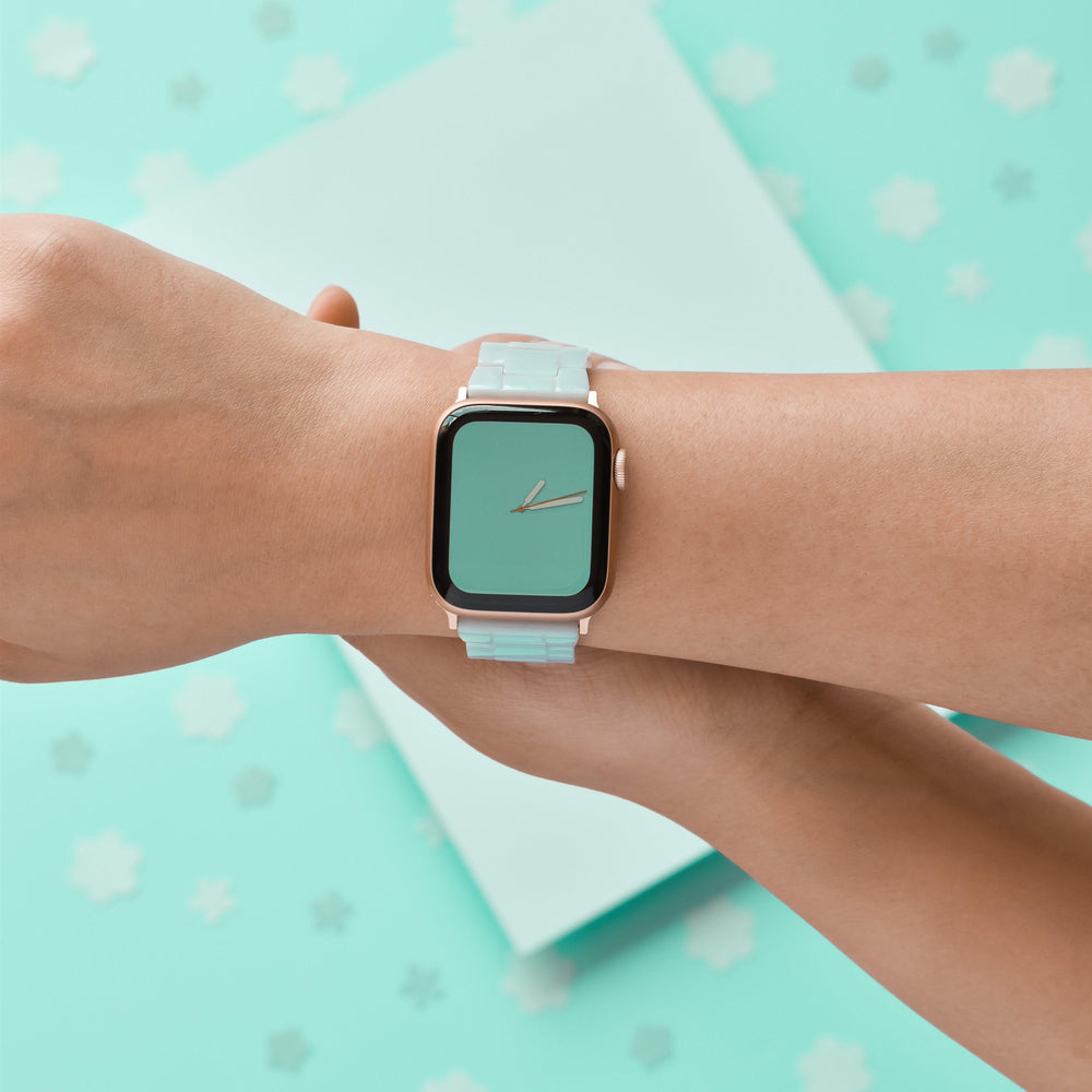 Close-up of a person's left wrist wearing a smart watch with Coconut Lane's Luxe Pastel Mint Apple Watch Strap. The watch face displays the time with tick marks and no numbers. The background features a matching light blue color with white star shapes dispersed around.