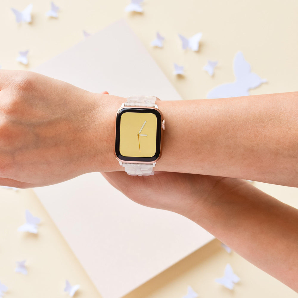 A person wearing a smartwatch with a minimalist single-hand clock design on a yellow display, paired with the Luxe Holo Butterfly Apple Watch Strap featuring a white wristband from Coconut Lane. The background is pale yellow with white paper butterflies scattered, creating an aesthetic and delicate scene. Holographic butterflies add sparkle to the scene.