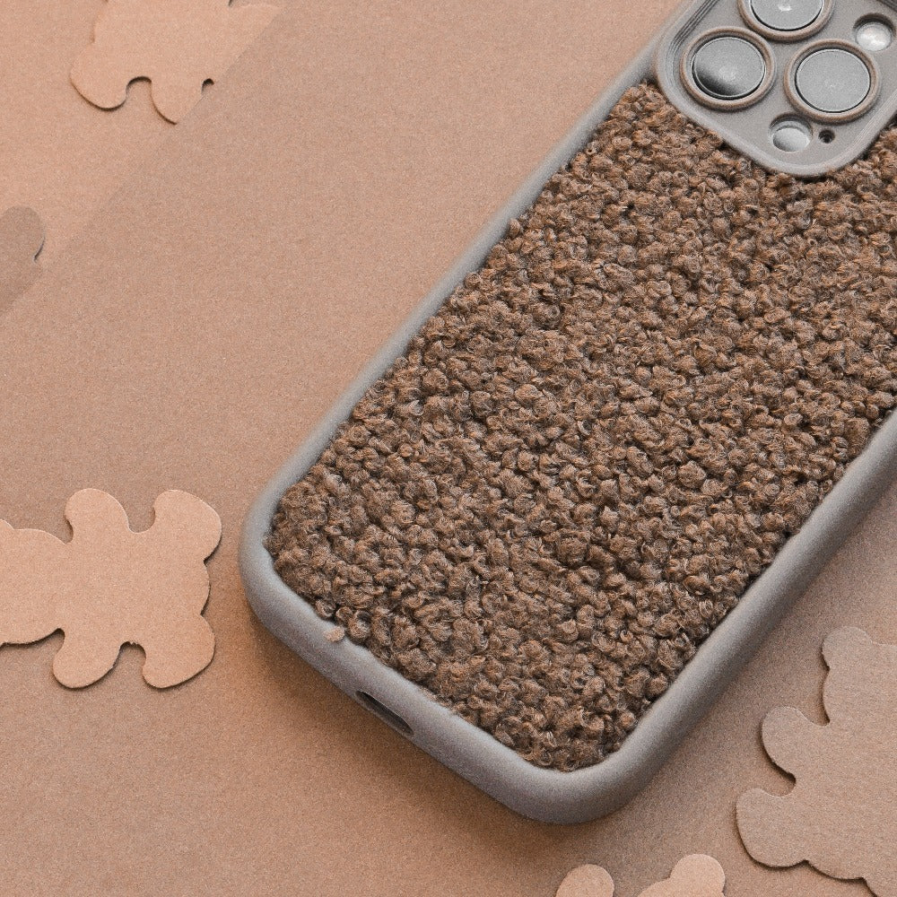 A close-up photo of a smartphone with a textured, brown, and rugged Cosy Teddy Phone Case - Brown by Coconut Lane resting on a surface scattered with various brown shapes. The phone's camera lenses are prominently visible at the top right corner.