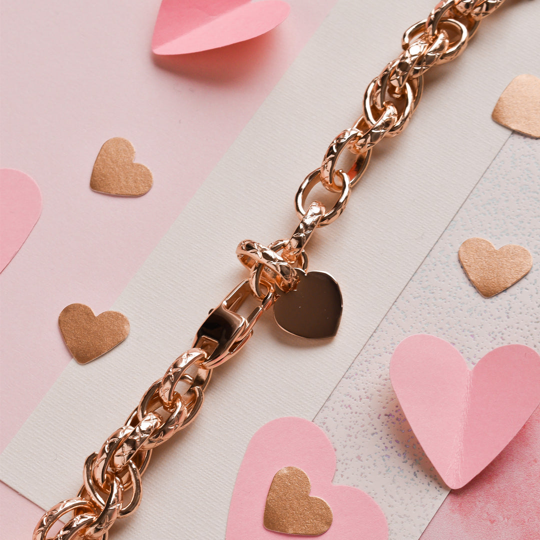 The Chain Bracelet Apple Watch Strap - Rose Gold by Coconut Lane, adorned with a heart-shaped charm, is surrounded by pink and gold paper hearts. The strap, featuring stainless steel metal accents, and the decorations are elegantly displayed on a light pink surface with some white and glitter accents.