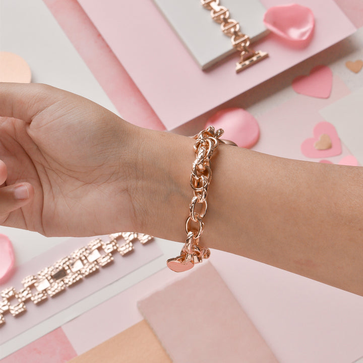 A hand adorned with a Coconut Lane Chain Bracelet Apple Watch Strap in Rose Gold, featuring heart charms, is displayed. The background includes scattered heart-shaped decorations and several pink and white rectangular items, enhancing the romantic and feminine ambiance of the scene.