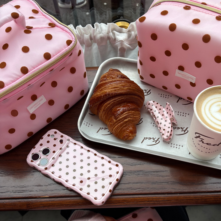flat lay picture in a cafe alongside a croissant and coffee. The phone shows the Dot Society pink and brown collection. Including a wavy pink and brown polka dot phone case, matching claw clip, vanity case and makeup bag. 