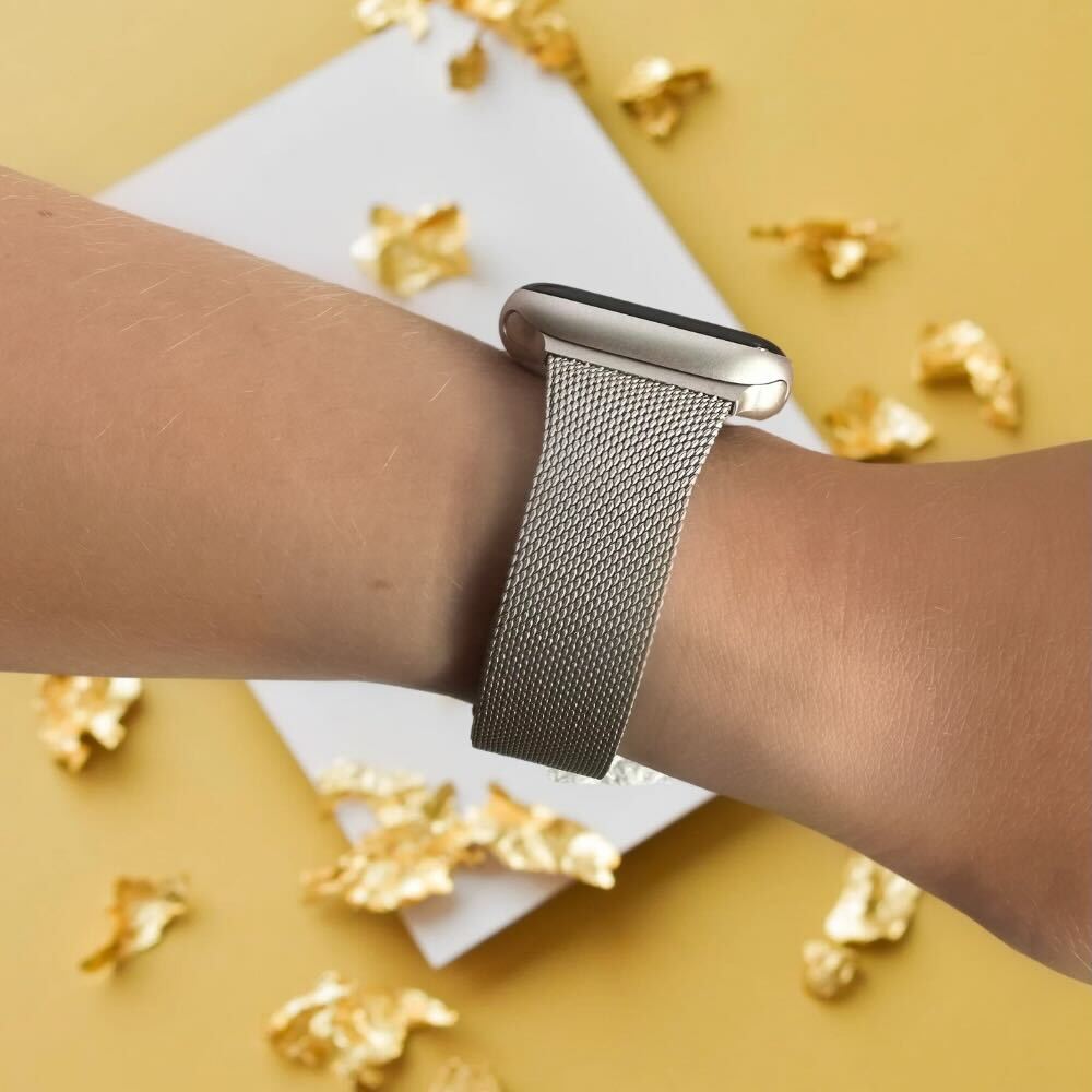 A close-up of a person's wrist wearing a Coconut Lane Milanese Apple Watch Strap - Starlight. The background consists of a yellow surface with scattered gold foil pieces, adding a touch of elegance to the minimalist composition.