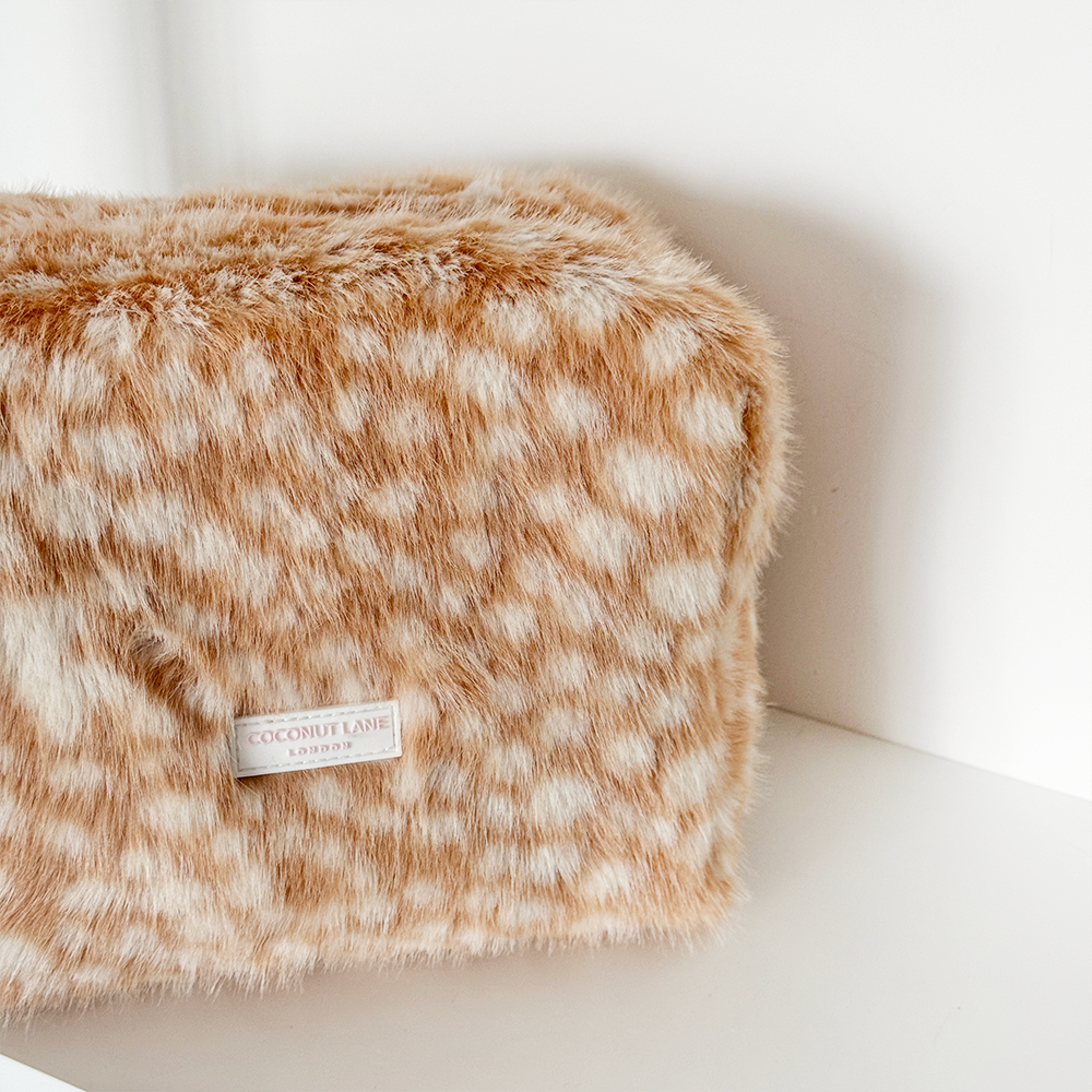 A close-up of a fluffy faux-fur cosmetic pouch in a warm tan and cream spotted pattern, with a visible Coconut Lane London label, resting on a white shelf against a white wall.