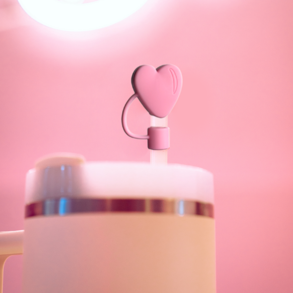 A close-up view of Coconut Lane's tumbler, featuring a pink heart-shaped lid decoration from their Pink Heart Straw Cover collection. The background is a soft pink, creating a cohesive and aesthetically pleasing effect. The flexible design includes a silicone straw cover for added convenience.