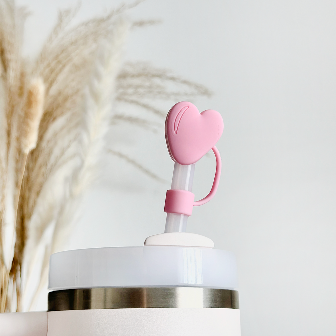 A white tumbler with a translucent straw is topped with Coconut Lane's Pink Heart Straw Cover, highlighting its flexible design. In the background, blurred pampas grass adds a touch of natural decor to the bright and minimalistic scene.