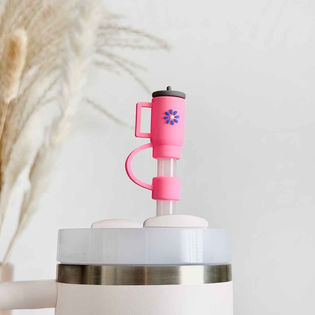 A close-up image of a white tumbler showcasing the Coconut Lane Mini Hot Pink Tumbler Straw Cover. The hygienic accessory features a blue and pink geometric flower design on the pink silicone cover. Blurred, light-colored dried plants are in the background against a white backdrop.