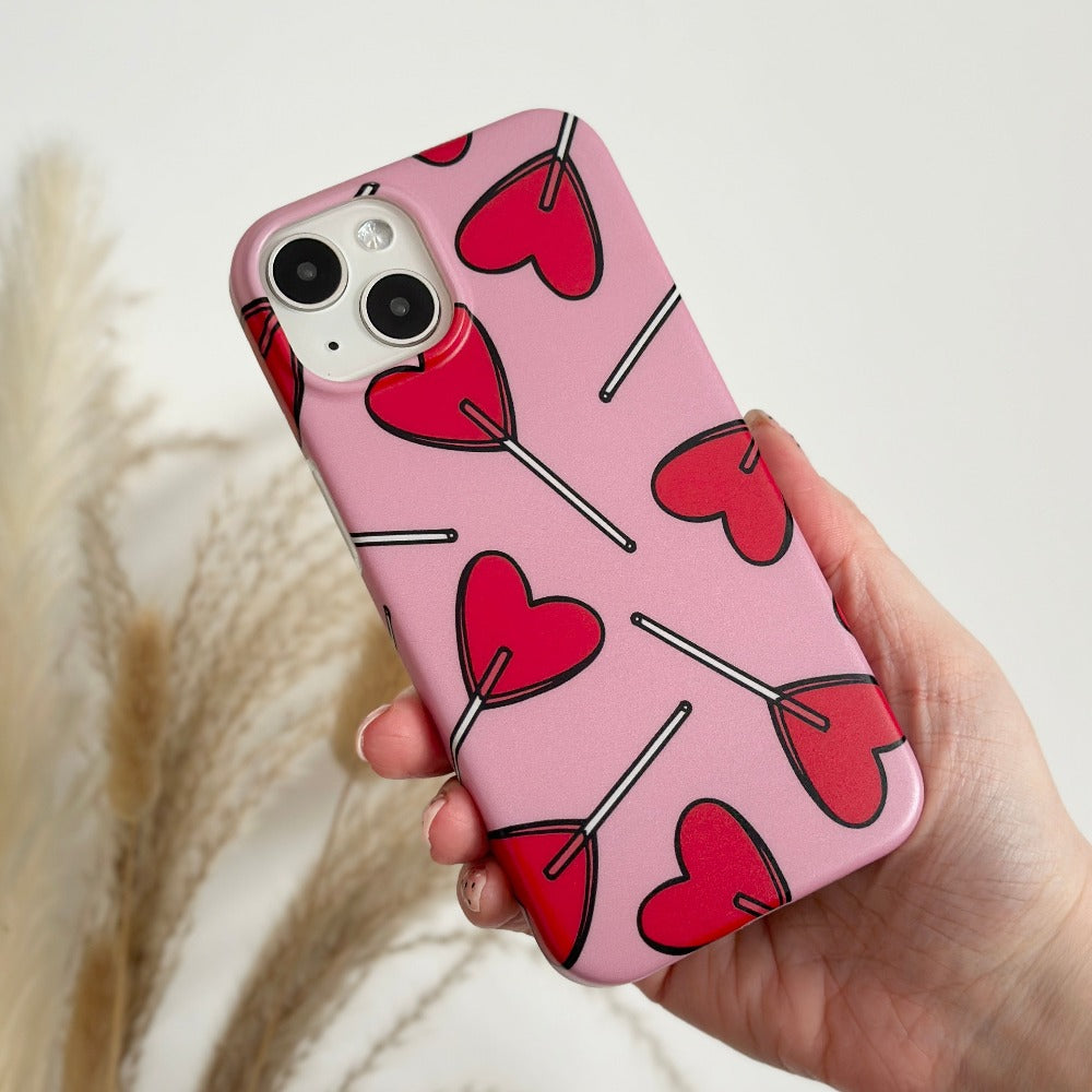 A hand holds the Coconut Lane Candy Hearts Phone Case, crafted from soft, flexible plastic and adorned with red heart lollipop illustrations against a light background featuring blurred, wispy plant elements.