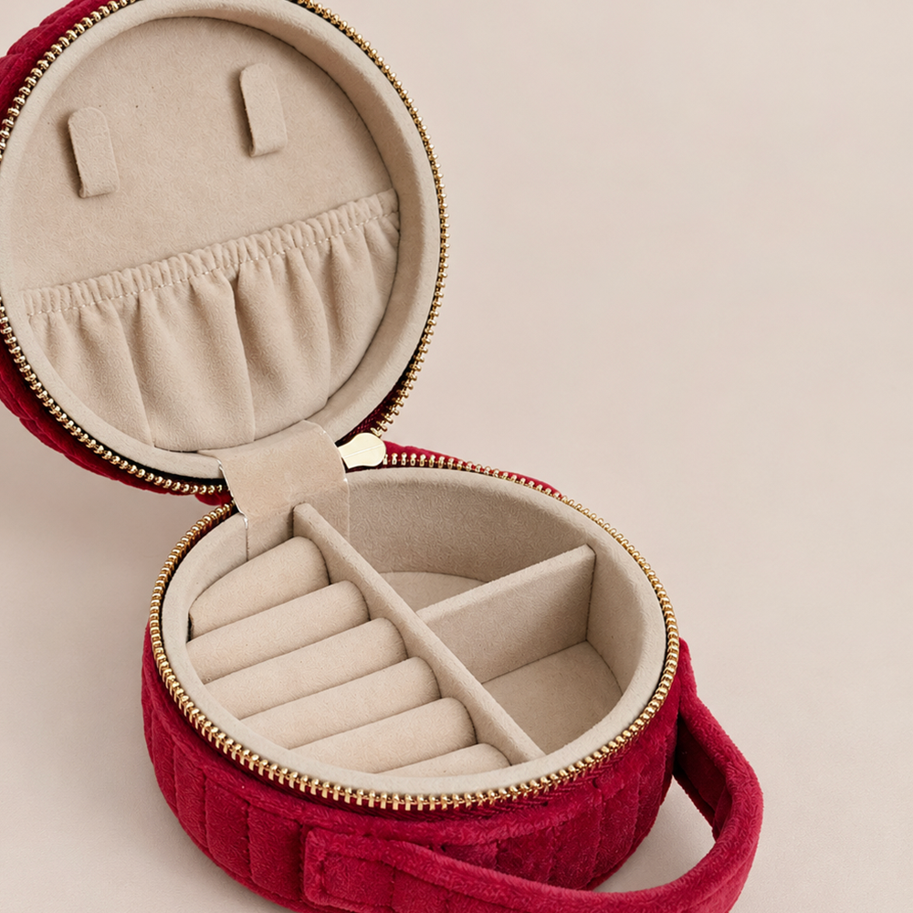 An open round red velvet jewelry organizer case with a beige fabric lining, gold-tone zipper, ring rolls, and a handle, shown on a light beige background.