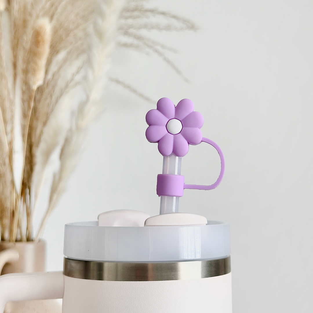 A close-up of a white tumbler with a silver rim, showcasing the Coconut Lane Lilac Flower Straw Cover. In the background, ornamental pampas grass stems are placed in a vase, slightly blurred to enhance the light and airy ambiance.