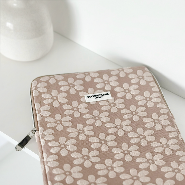 A pink and white floral-patterned laptop case with a zipper, labeled "Coconut Lane," lies on a white surface next to a white vase. This elegant Mocha Flower iPad Sleeve can also be used for your laptop.