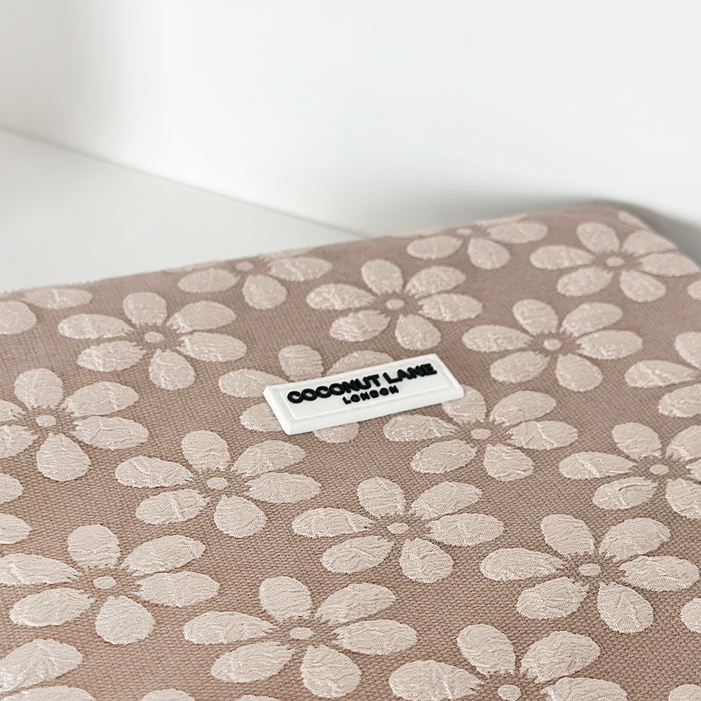 A Mocha Flower iPad Sleeve by Coconut Lane is shown, featuring a beige background adorned with a pattern of white flowers. A small, centered tag reads "Coconut Lane London.