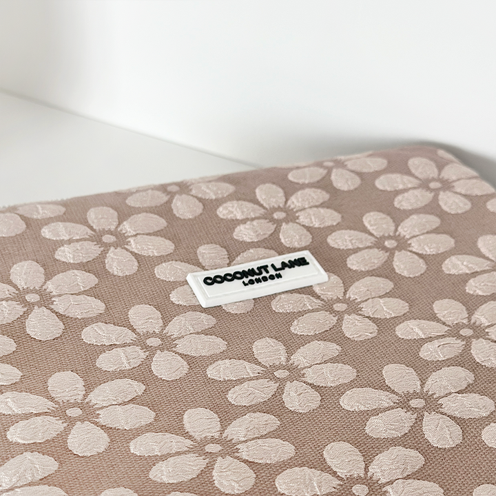 A Mocha Flower iPad Sleeve by Coconut Lane is shown, featuring a beige background adorned with a pattern of white flowers. A small, centered tag reads "Coconut Lane London.