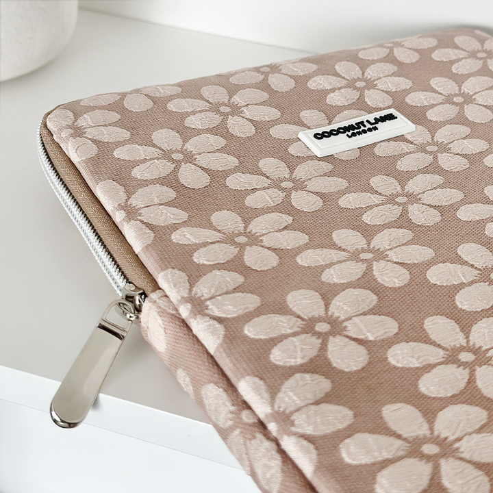 A close-up of a pink Mocha Flower iPad Sleeve with white floral patterns and a silver zipper. The sleeve, resembling an elegant embroidered pouch, has a white label that reads, "COCONUT LANE London." It is placed on a white surface.
