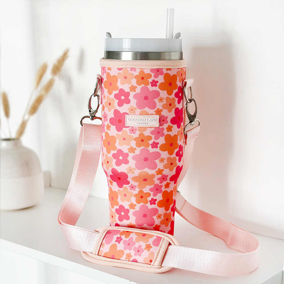 A reusable drink cup with a straw is nestled in Coconut Lane's Tumbler Carry Case - Pink & Orange Bloom, featuring vibrant floral fabric in shades of pink, red, and orange. The case includes a light pink shoulder strap for effortless carrying. Positioned on a white surface, the travel mug is beautifully complemented by dried grasses in a vase in the background.