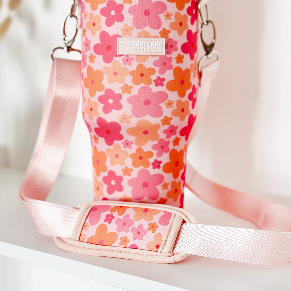 A pink strap with a shoulder pad is attached to a vibrant insulated bottle holder adorned with a pattern of pink, peach, and orange flowers. The fabric label on the holder reads "Coconut Lane." Perfect as the Tumbler Carry Case - Pink & Orange Bloom for your travel mug or Stanley Quencher, this item is displayed on a white surface.