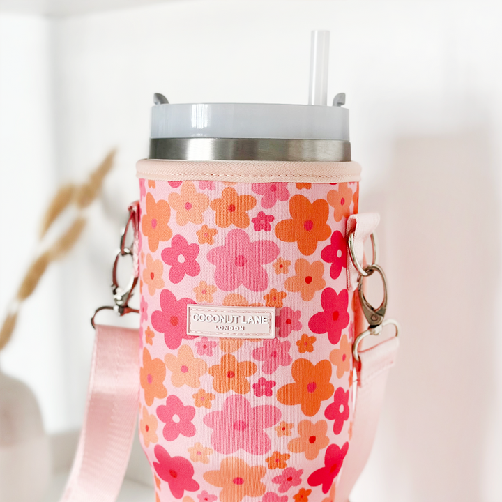 A tall drinking cup with a straw, encased in the "Tumbler Carry Case - Pink & Orange Bloom" by Coconut Lane. The background features blurred neutral tones and visible dried plants.