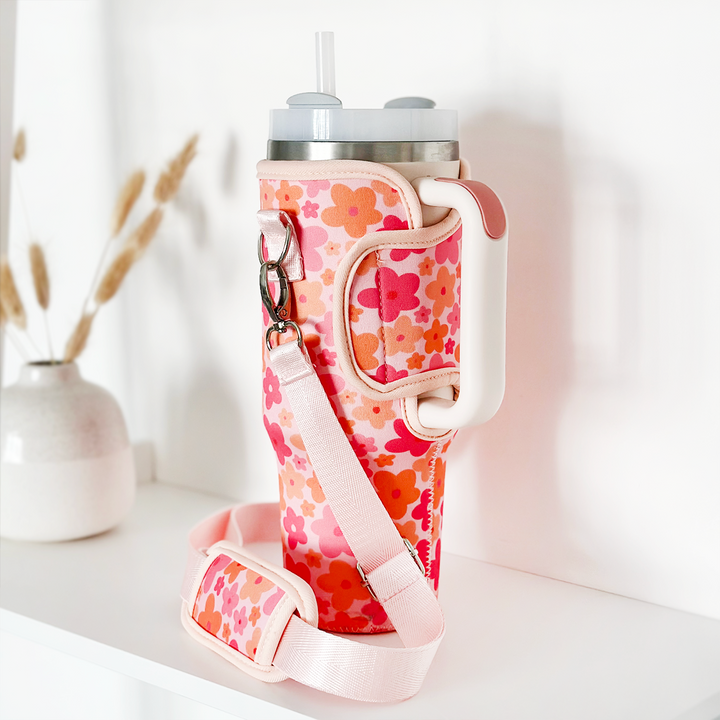 A Stanley Quencher with a spout lid is nestled in a Coconut Lane Tumbler Carry Case - Pink & Orange Bloom, featuring a handle and adjustable strap, on a white surface. In the background, there's a white vase containing some dried grasses.