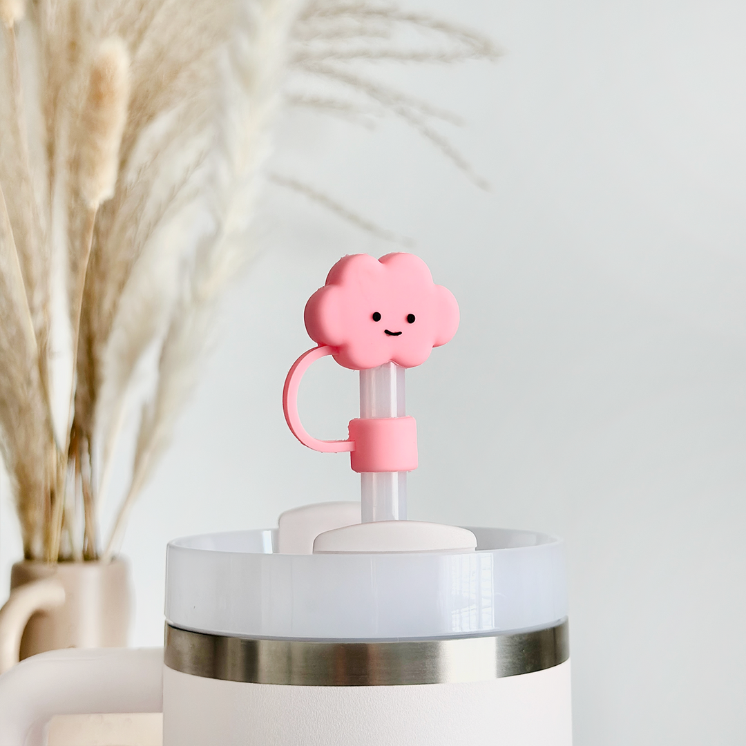 The Pink Cloud Straw Cover by Coconut Lane features a pink, cloud-shaped silicone cover with a smiling face that adorns the straw of a white tumbler with a metal ring around the top. In the background, dried pampas grass in a vase is slightly out of focus against a light-colored wall.