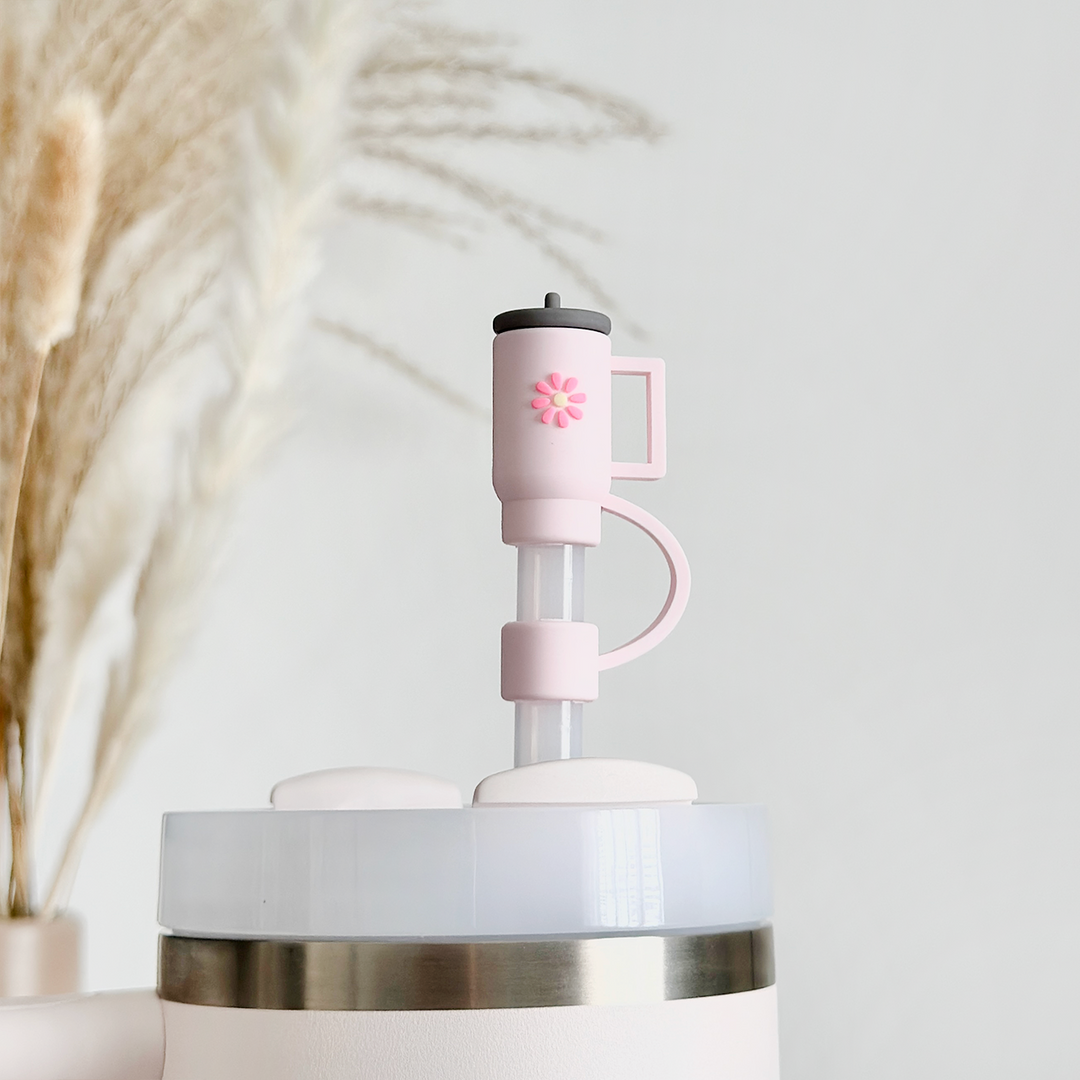 A close-up of the Coconut Lane Mini Pink Tumbler Straw Cover, showcasing the light pink portable coffee press attached to the top of a travel mug. The press features a handle and a plunger with an elegant floral design, complemented by a silicone straw cover. In the blurred background, beige pampas grass adds a decorative touch.